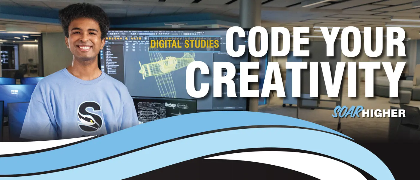 Brandon Dyson in a computer lab wearing a light blue SOAR HIGHER t‑shirt stands in front of monitors displaying a 3D airplane model and technical drawings, representing digital design and coding work. Text on the image says - Code Your Creativity.