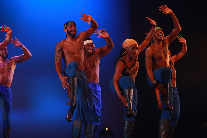 A group of dancers with their arms raised in front of a blue-lit backdrop