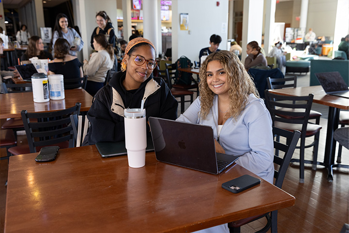 Two female students smile while sitting at a table in the Campus Center Coffeehouse with a laptop, cell phone, and water bottle on the table