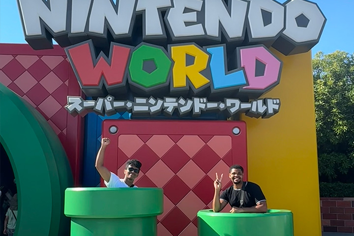 A Nintendo World sign, Japanese characters, and two students posing in large green tubes from Mario World