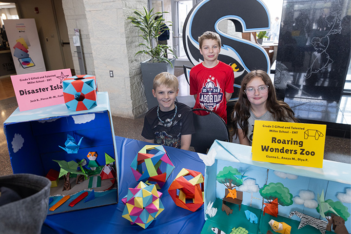 Three elementary school children sit with their origami works on display in the Campus Center