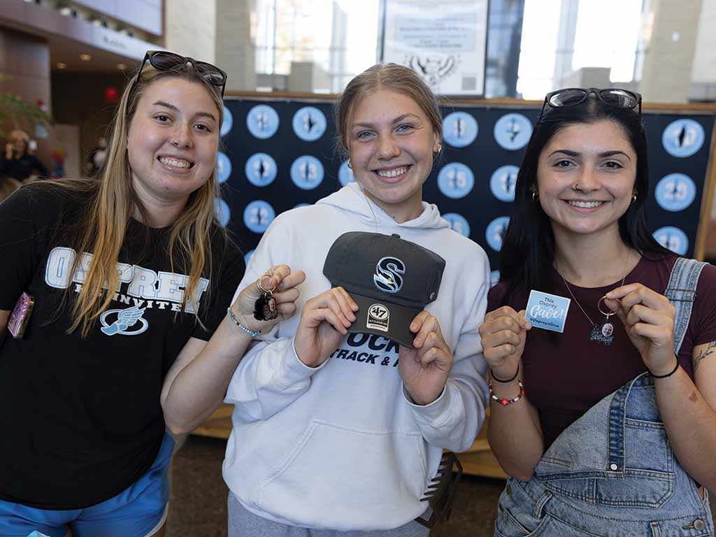 Three female students hold up prizes from the punch board, including keychains and a Stockton hat