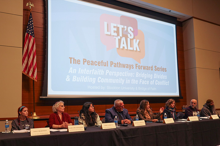 Panelists sit behind a long table with a screen behind them announcing the name of the event 'Let's Talk'