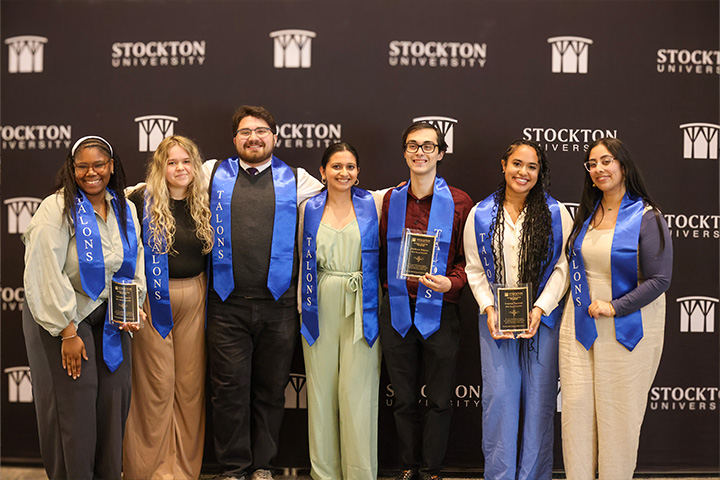 A group of Stockton students stand in front of a black step-and-repeat banner with the Stockton logo, while wearing special blue honor stoles and holding award plaques