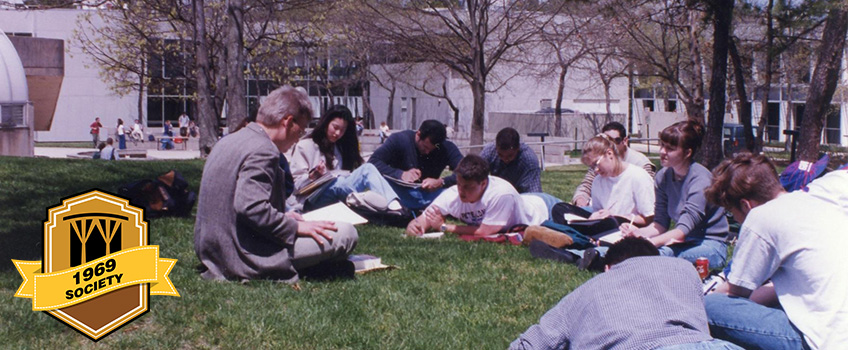 Photo from the early days on campus, students sitting in a circle on the quad and listening to their lecturer