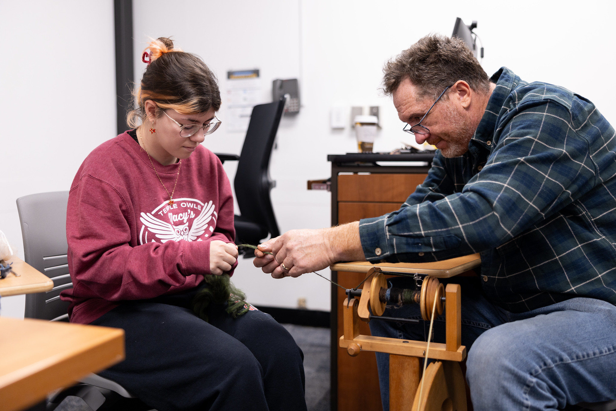A woman in a red shirt holds the end of a string while a man in a blue, plaid shirt helps spool thread through a wooden machine.