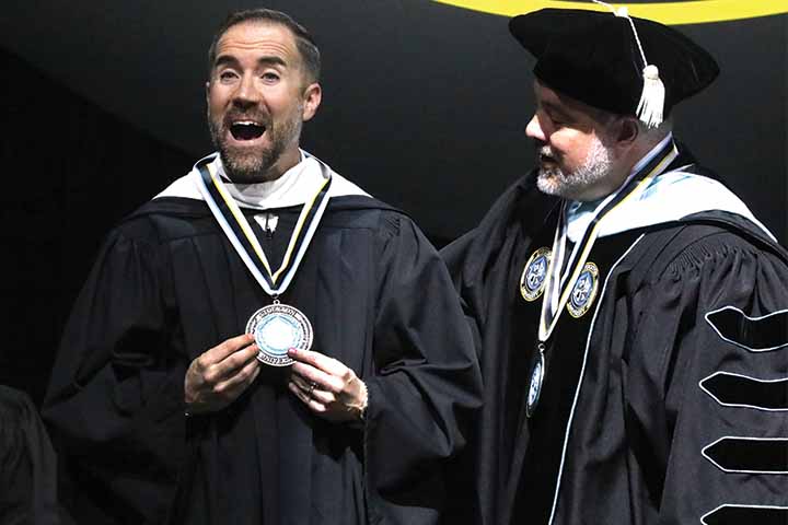 Dave Sholler in graduation gown excitedly receives the Presidential Medal of Distinction from Joe Bertolino